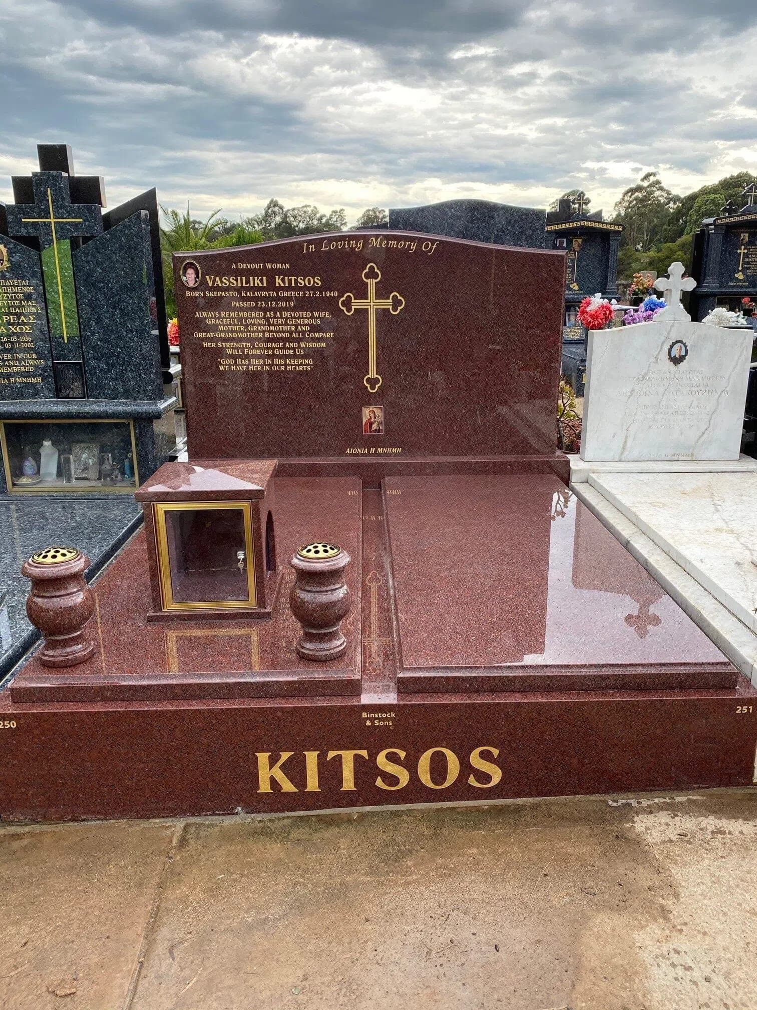 Double full Greek monument with a serpentine-shaped headstone, gold leaf lettering, ruby red granite, and a candle box.